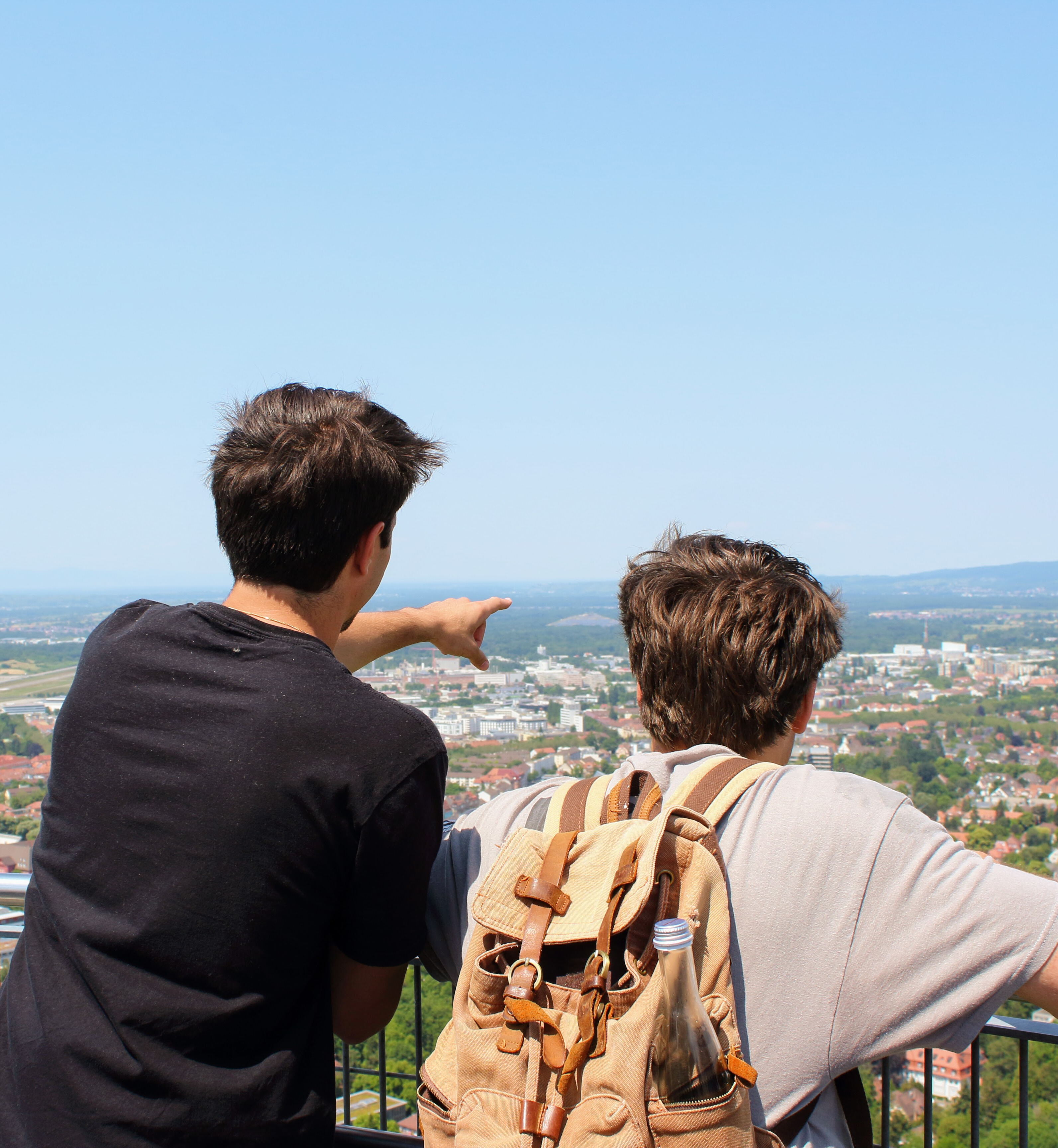 Two young men stand with their backs to the camera, leaning against a railing and looking out over a view of green trees and orange-roofed German buildings. One of the men points to something in the distance. The sky is blue and cloudless, and hazy mountains can be seen in the distance. 