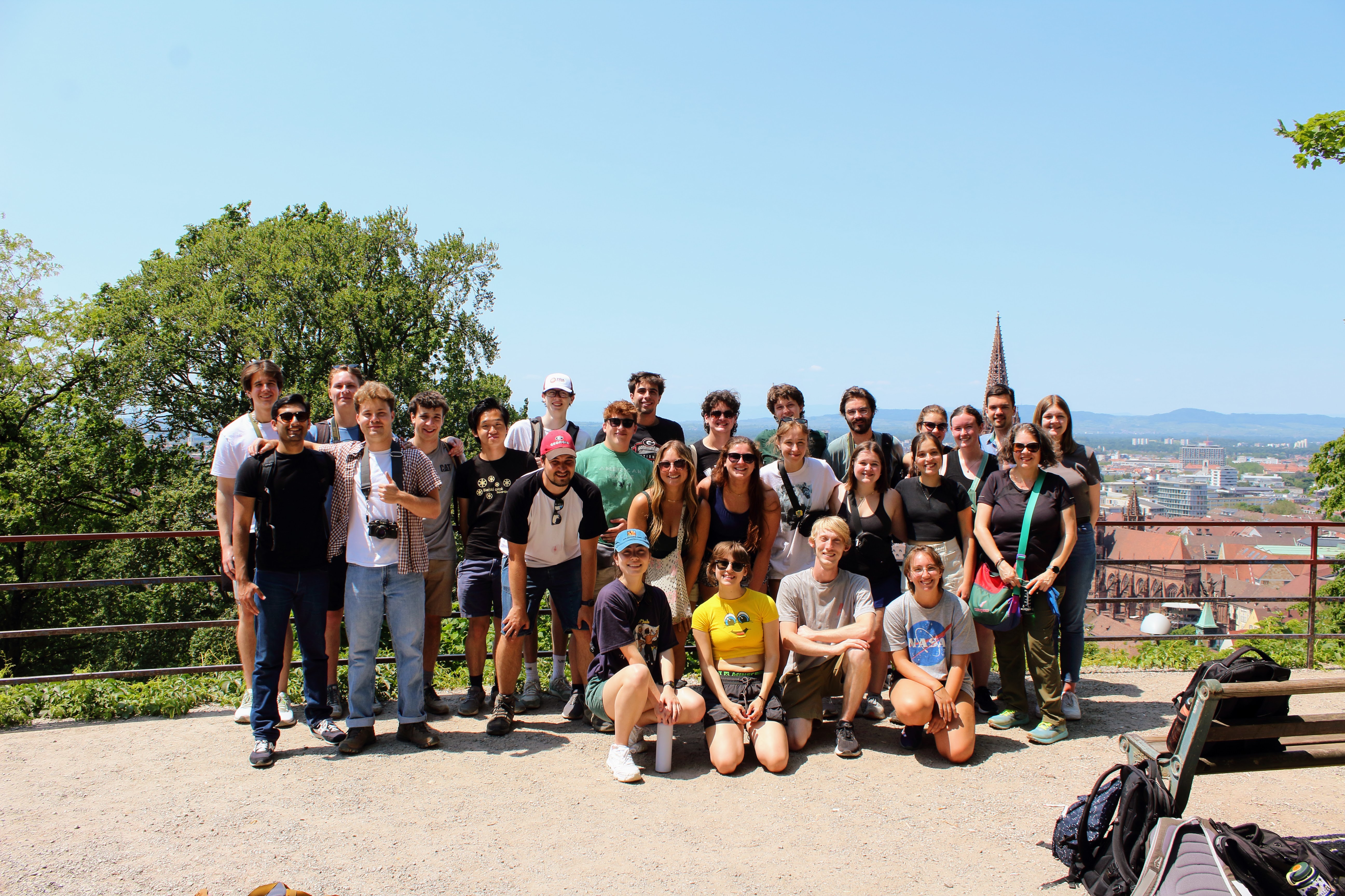 Several students stand in front of an overlook. A German city, with orange roofs and a tall spire, can be seen in the background, behind which are mountains. The sky is blue, and the sun is shining. The students are all smiling.
