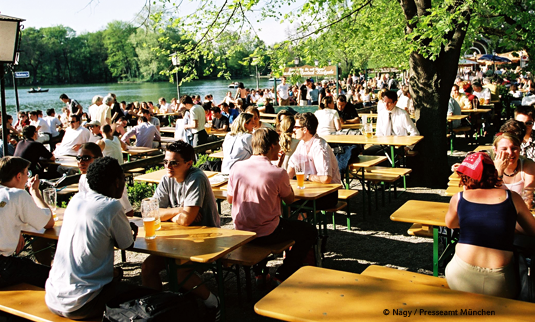 Beer garden in Munich with people seated next to a river enjoying a warm day.