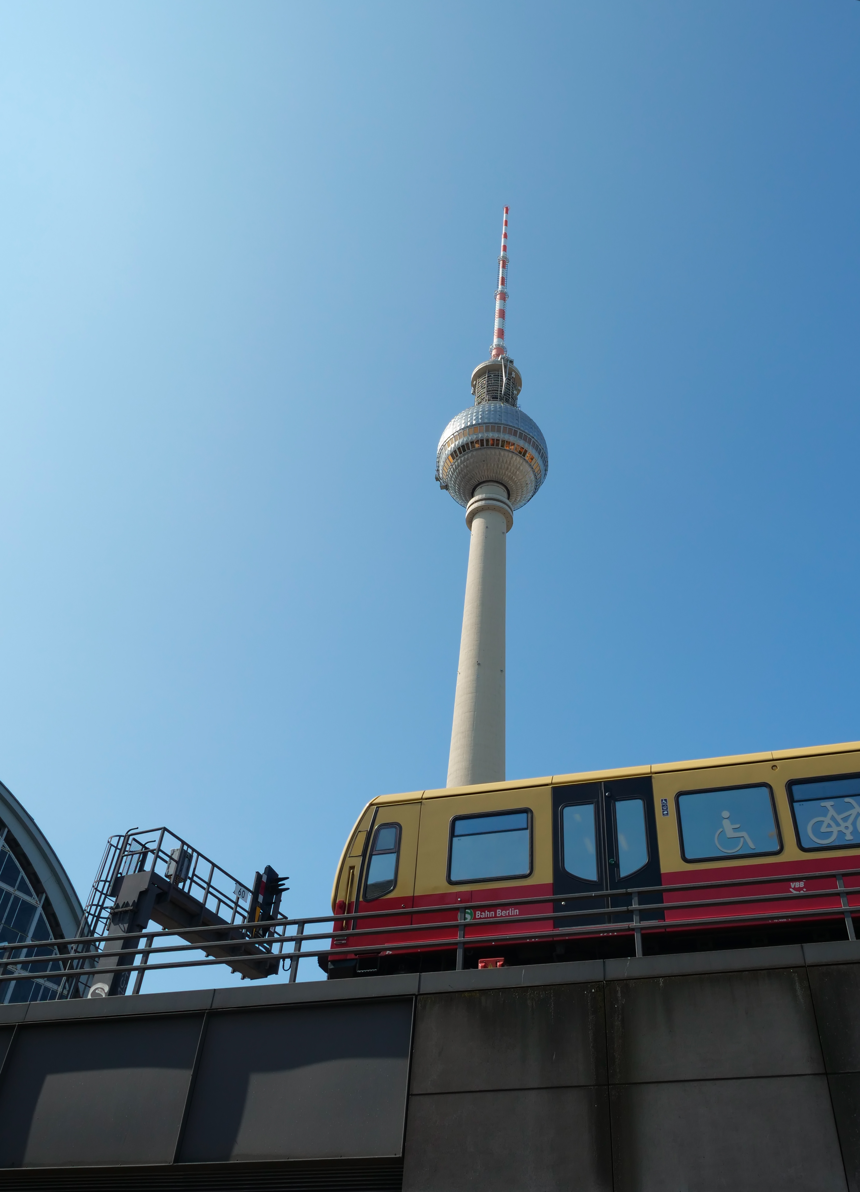 A yellow and red train passes in front of the Berlin Fernsehturm, or television tower, a tall, white needle-like structure.